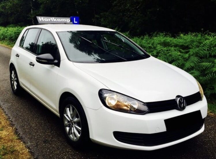 De witte Volkswagen Golf TDI lesauto van Autorijschool Hartkamp met daklicht op een bosweg bij Barneveld